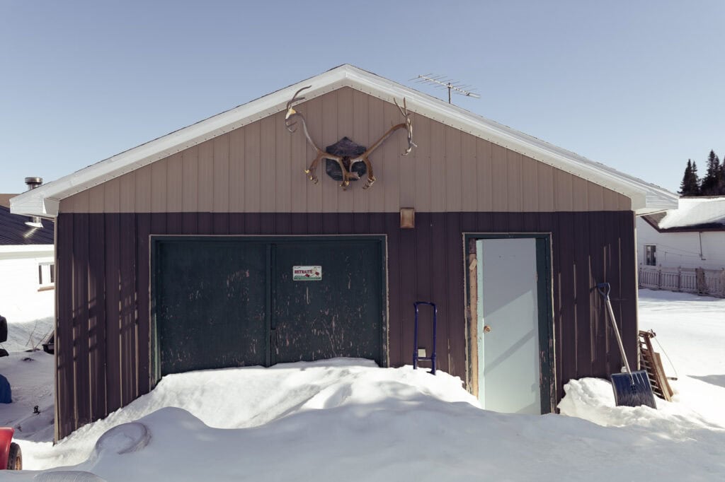 Garage brun à deux portes partiellement ensevelies sous la neige, avec un grand panache d’orignal accroché au centre de la façade, à Tête-à-la-Baleine.