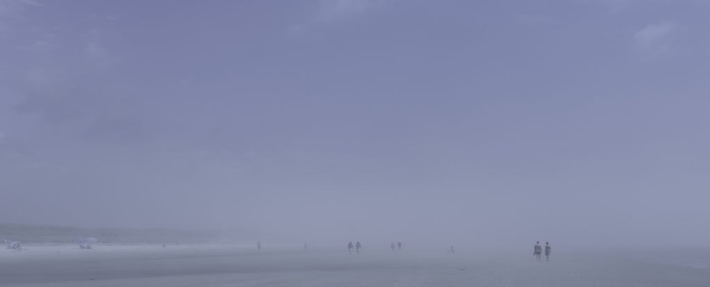 Plage panoramique dans un brouillard bleu dense. À droite, un couple marche au bord de l’eau, tandis qu’à gauche quelques parasols et d’autres figures apparaissent dans la brume.