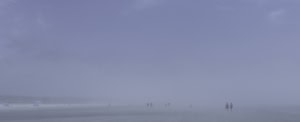 Plage panoramique dans un brouillard bleu dense. À droite, un couple marche au bord de l’eau, tandis qu’à gauche quelques parasols et d’autres figures apparaissent dans la brume.
