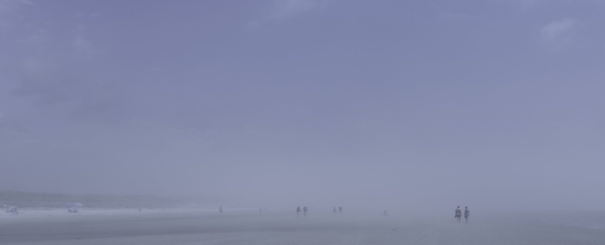 Plage panoramique dans un brouillard bleu dense. À droite, un couple marche au bord de l’eau, tandis qu’à gauche quelques parasols et d’autres figures apparaissent dans la brume.