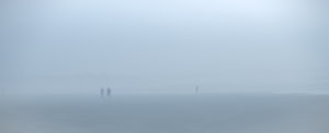 Une plage panoramique, lisse et uniforme, s’étend dans un brouillard bleu assez dense jusqu’à une ligne d’arbres à peine visible. Le ciel occupe un peu plus de la moitié supérieure de l’image. À gauche, un couple marche vers nous. Plus loin, une silhouette solitaire semble aller dans l’autre sens dans la brume.