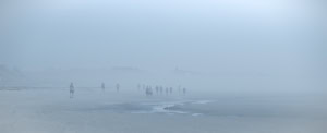 Plage panoramique dans un brouillard bleu dense, avec une mare résiduelle au premier plan et plusieurs promeneurs avançant sur le rivage.