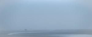Plage panoramique dans un brouillard bleu dense. Des mares résiduelles apparaissent sur le sable, un couple se distingue au loin, suivi par d’autres silhouettes.