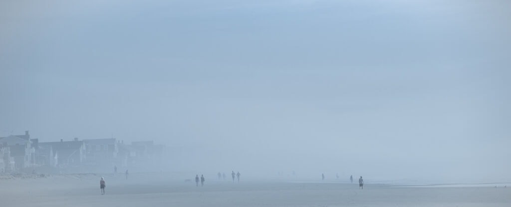 Plage panoramique dans un brouillard bleu dense. À gauche, des maisons bordent le rivage, tandis que plusieurs silhouettes avancent dans la brume.