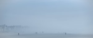 Plage panoramique dans un brouillard bleu dense. À gauche, des maisons bordent le rivage, tandis que plusieurs silhouettes avancent dans la brume.