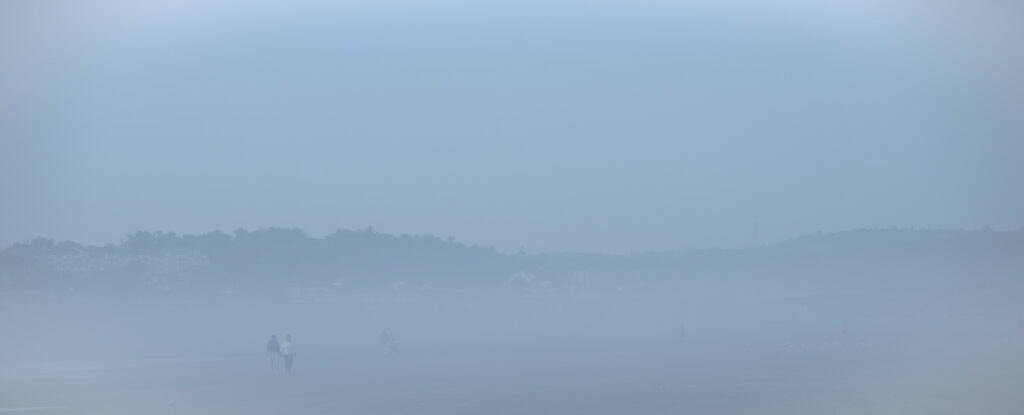 Plage panoramique dans un brouillard bleu dense, limitée par une colline arborée. À gauche, près de l’eau, un couple se détache dans la brume.