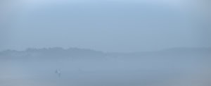 Plage panoramique dans un brouillard bleu dense, limitée par une colline arborée. À gauche, près de l’eau, un couple se détache dans la brume.