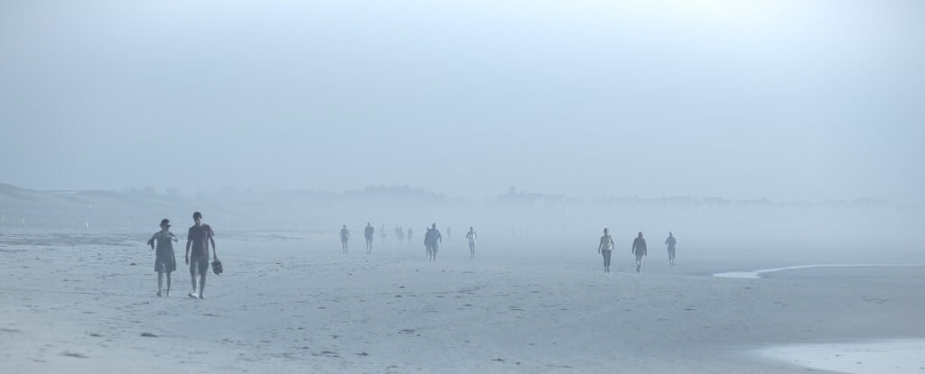 Plage panoramique dans un brouillard bleu dense. Plusieurs marcheurs avancent vers le photographe, dont un couple à gauche semblant discuter.