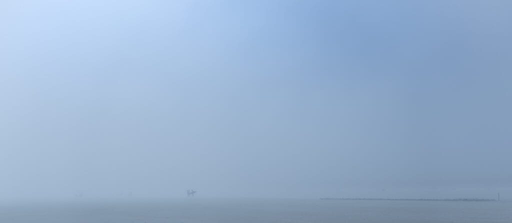 Plage panoramique dans un brouillard bleu dense. Très loin, deux personnes reviennent de la baignade, dont l’une porte une planche de surf. À droite, une fine ligne rocheuse semble marquer la limite entre le sable et l’eau.