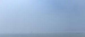 Plage panoramique dans un brouillard bleu dense. Très loin, deux personnes reviennent de la baignade, dont l’une porte une planche de surf. À droite, une fine ligne rocheuse semble marquer la limite entre le sable et l’eau.