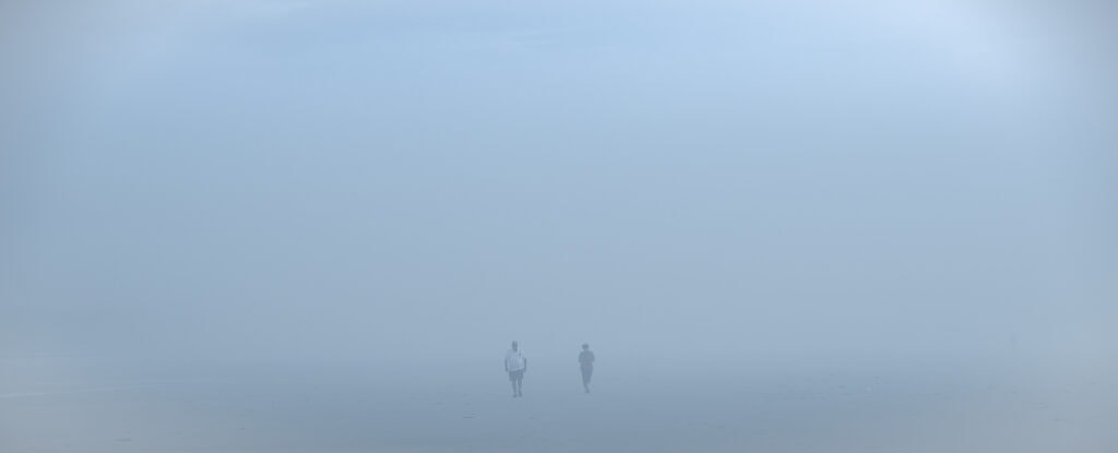 Plage panoramique dans un brouillard bleu très dense. Un homme en polo blanc marche vers le photographe et croise une femme.