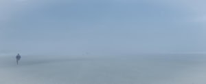 Plage panoramique dans un brouillard bleu dense. À gauche, un coureur s’éloigne, tandis que quelques silhouettes apparaissent très loin dans la brume.