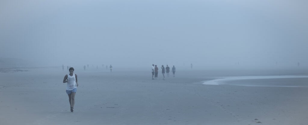 Plage panoramique dans un brouillard bleu dense. À gauche, une joggeuse court vers nous, tandis qu’au centre un petit groupe discute et que d’autres silhouettes apparaissent plus loin.