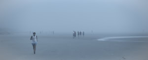 Plage panoramique dans un brouillard bleu dense. À gauche, une joggeuse court vers nous, tandis qu’au centre un petit groupe discute et que d’autres silhouettes apparaissent plus loin.