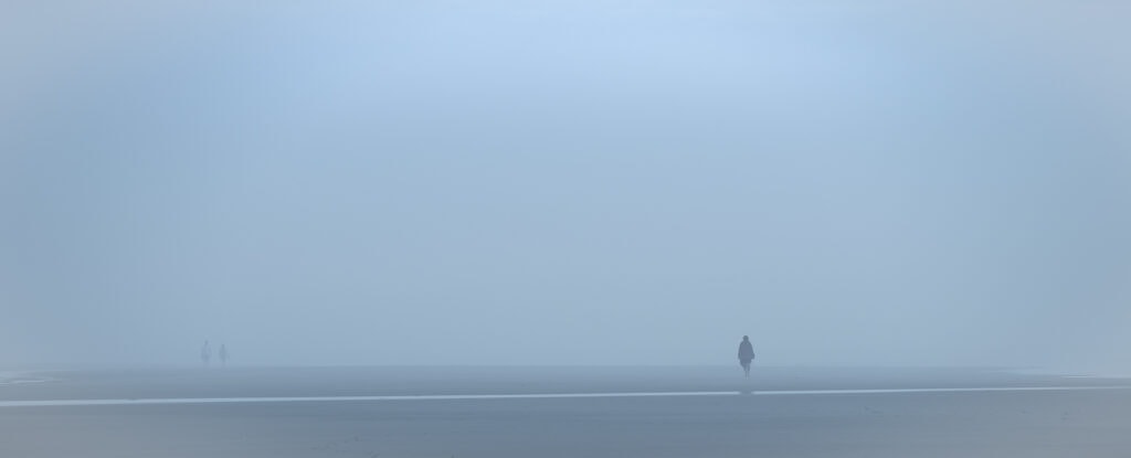 Plage panoramique dans un brouillard bleu dense. Une silhouette marche seule à droite, tandis que deux autres apparaissent très loin à gauche.