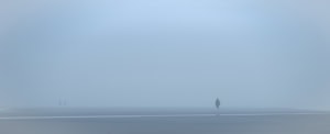 Plage panoramique dans un brouillard bleu dense. Une silhouette marche seule à droite, tandis que deux autres apparaissent très loin à gauche.