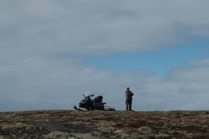 Homme debout près d’une motoneige au sommet d’un terrain rocheux couvert de mousses et de lichens, sur l’île Anderson, à Tête-à-la-Baleine.