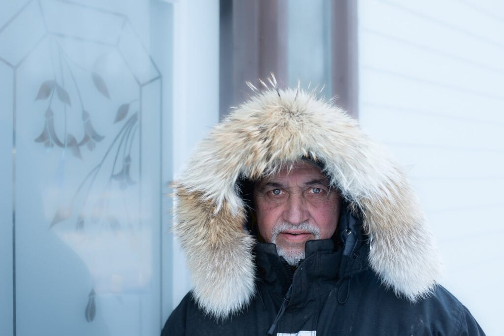 Portrait de face d’un homme grisonnant en anorak foncé à capuchon bordé de fourrure, devant le mur d’une maison, à Tête-à-la-Baleine.