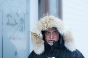 Portrait de face d’un homme grisonnant en anorak foncé à capuchon bordé de fourrure, devant le mur d’une maison, à Tête-à-la-Baleine.