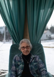 Femme aux cheveux gris assise dans un fauteuil vert devant de grandes fenêtres encadrées de rideaux verts, à Tête-à-la-Baleine.