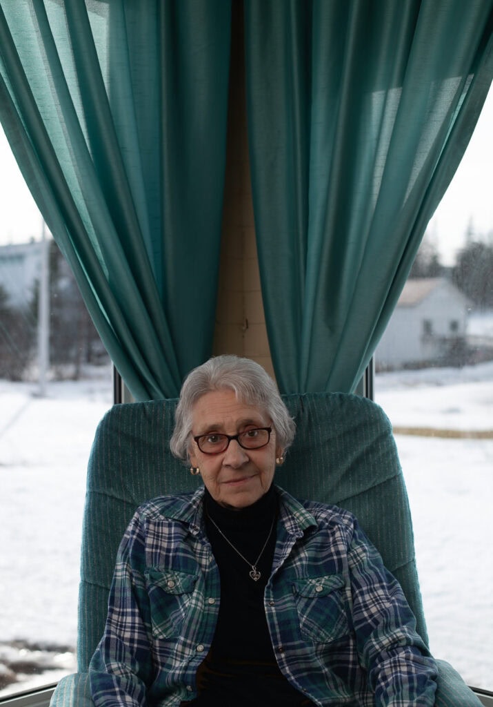 Femme aux cheveux gris assise dans un fauteuil vert devant de grandes fenêtres encadrées de rideaux verts, à Tête-à-la-Baleine.
