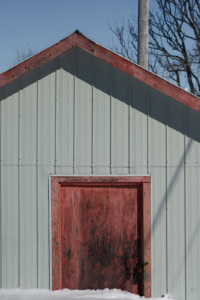Façade de remise en tôle grise avec porte rouge usée partiellement recouverte de neige, à Tête-à-la-Baleine.