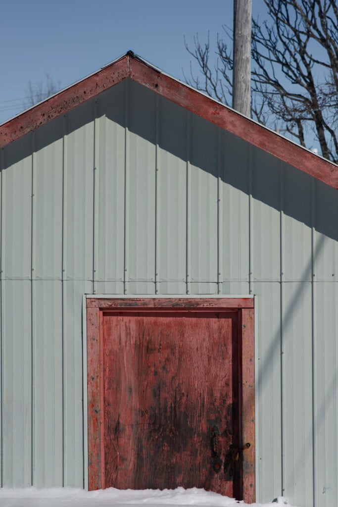Façade de remise en tôle grise avec porte rouge usée partiellement recouverte de neige, à Tête-à-la-Baleine.