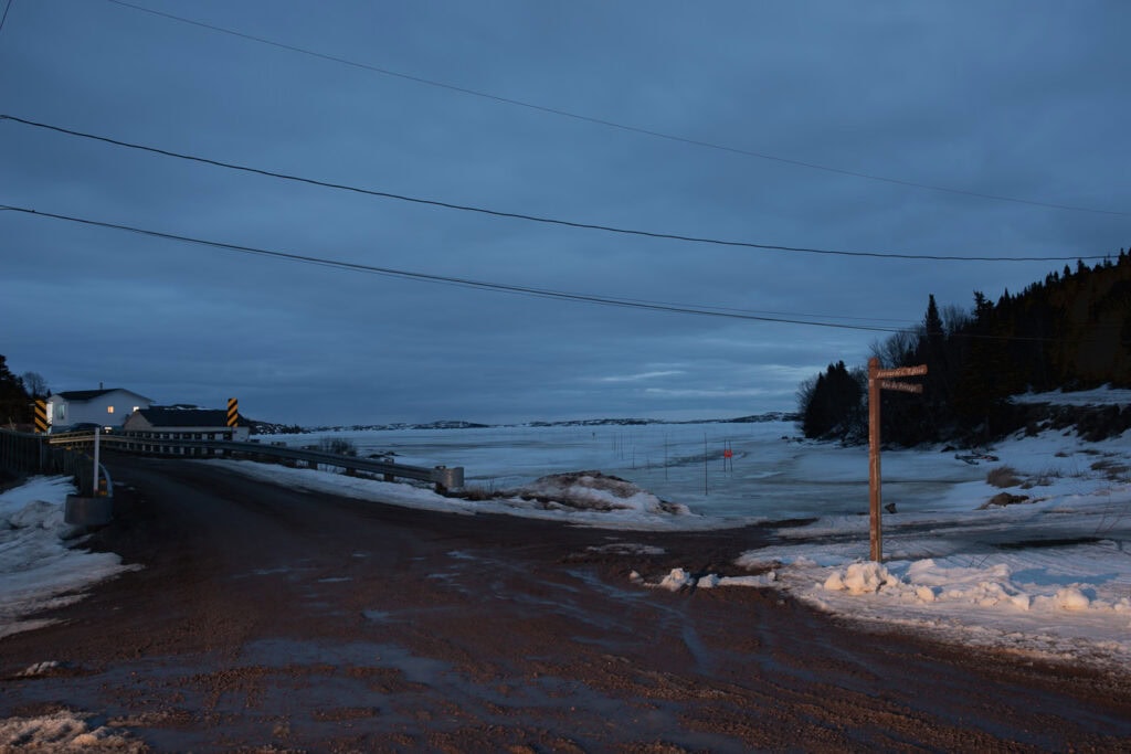 Route boueuse en fin d’hiver menant à un pont, devant une baie gelée où l’on distingue le balisage de la Route blanche, à Tête-à-la-Baleine.