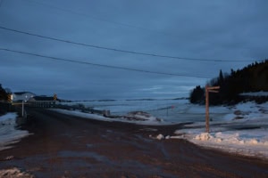 Route boueuse en fin d’hiver menant à un pont, devant une baie gelée où l’on distingue le balisage de la Route blanche, à Tête-à-la-Baleine.