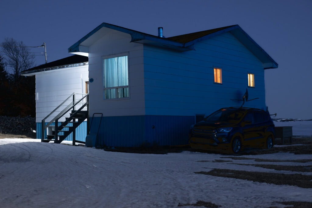 Maison isolée, de nuit en hiver, vue de trois-quarts, avec fenêtres éclairées, escalier extérieur et voiture stationnée sur le côté, à Tête-à-la-Baleine.