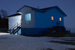 Maison isolée, de nuit en hiver, vue de trois-quarts, avec fenêtres éclairées, escalier extérieur et voiture stationnée sur le côté, à Tête-à-la-Baleine.