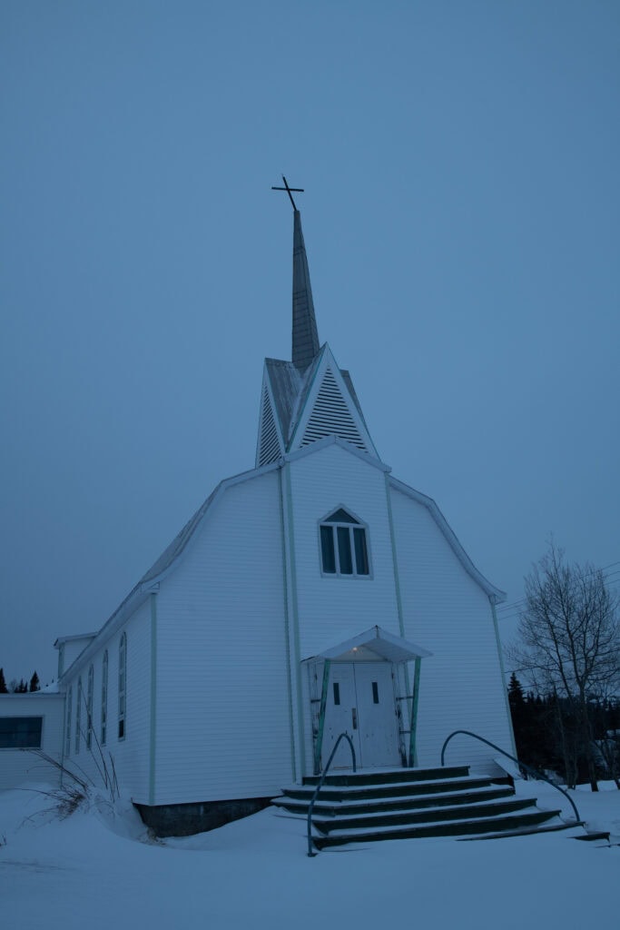 Façade d’église vue en léger oblique, de nuit, entourée de neige, avec escalier, double porte et clocher surmonté d’une croix penchée, à Tête-à-la-Baleine.