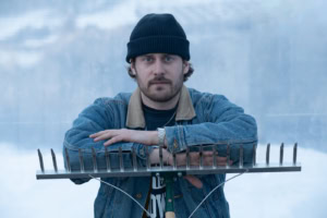 Portrait en buste d’un jeune homme barbu portant un bonnet foncé et une veste en denim, accoudé sur un râteau retourné, à Tête-à-la-Baleine.