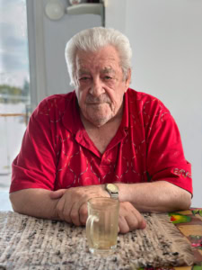 Portrait de face d’un homme âgé en chemise rouge, accoudé à une table avec un verre contenant un fond de café, à Tête-à-la-Baleine.