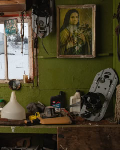 Coin de garage peint en vert avec étagère encombrée d’objets, fenêtre, image pieuse et raquette à neige, à Tête-à-la-Baleine.