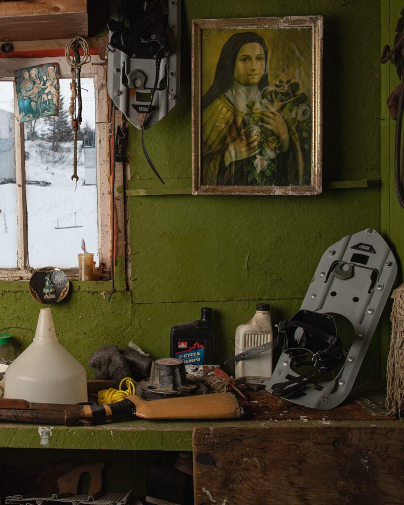 Coin de garage peint en vert avec étagère encombrée d’objets, fenêtre, image pieuse et raquette à neige, à Tête-à-la-Baleine.