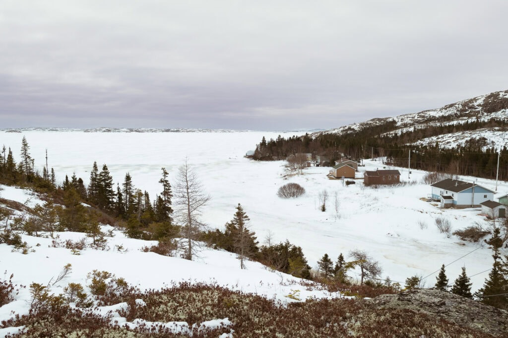 Paysage enneigé à la fin de l’hiver avec rivière gelée, maisons, collines boisées et étendue glacée, à Tête-à-la-Baleine.