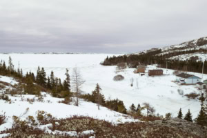 Paysage enneigé à la fin de l’hiver avec rivière gelée, maisons, collines boisées et étendue glacée, à Tête-à-la-Baleine.