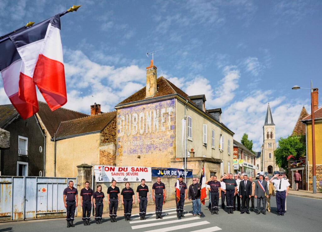 Pompiers au garde-à-vous avec le maire et des élus lors d’un instant de recueillement sur une place de Sainte-Sévère-sur-Indre, avec drapeaux français et église au loin.