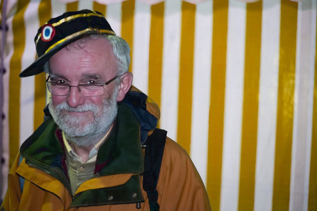 Portrait d’un homme barbu portant de travers un képi de facteur devant une tente rayée blanche et ocre à Sainte-Sévère-sur-Indre.
