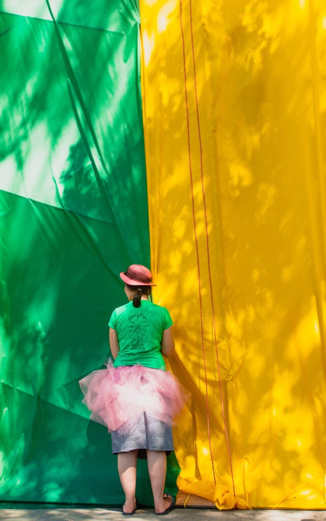 Jeune femme vue de dos devant deux grandes tentures verte et jaune, portant un chapeau rouge, un t-shirt vert et un tutu rose à Saint-Élie-de-Caxton.