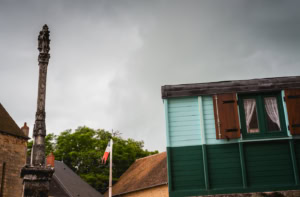 Place de village en contreplongée avec calvaire de pierre, drapeau tricolore et roulotte de forains sous un ciel gris, à Sainte-Sévère-sur-Indre.