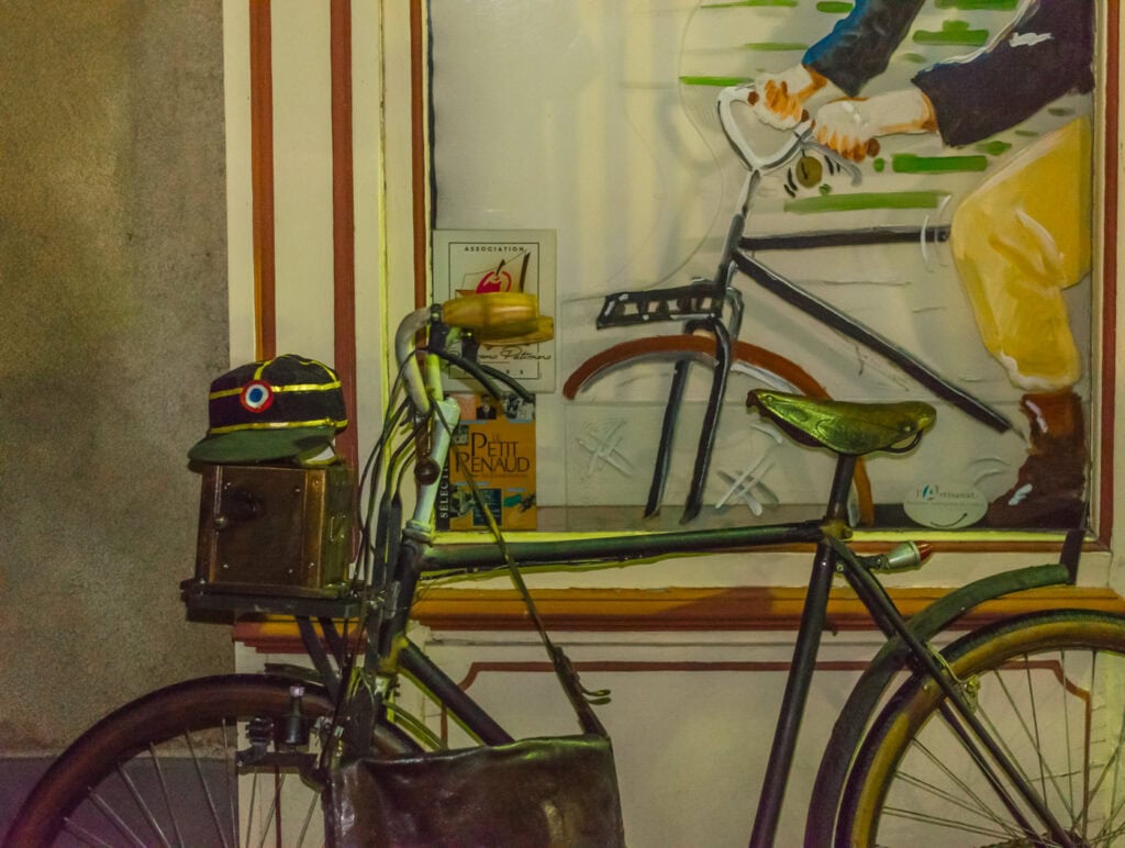 Réplique du vélo du facteur de Jour de fête adossée à une vitrine peinte, avec képi et sacoche, de nuit à Sainte-Sévère-sur-Indre.