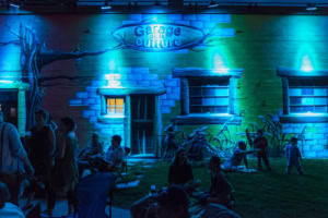 Rassemblement nocturne devant la façade peinte du Garage de la culture, éclairée en bleu et vert, avec adultes assis et enfants jouant sur la pelouse à Saint-Élie-de-Caxton.