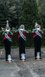 Trois trompettistes de fanfare alignés devant la haie d’un cimetière, le visage caché par le pavillon de leur instrument, avec leur képi à plumes posé à leurs pieds.