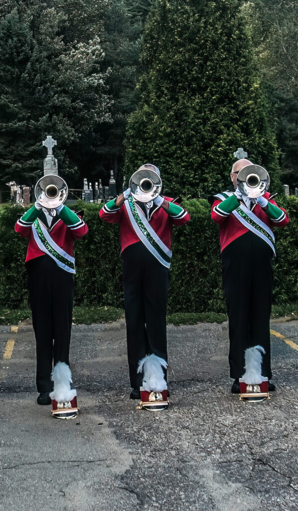 Trois trompettistes de fanfare alignés devant la haie d’un cimetière, le visage caché par le pavillon de leur instrument, avec leur képi à plumes posé à leurs pieds.