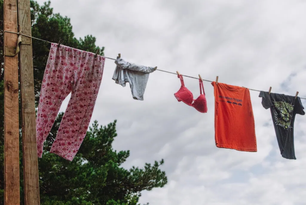Photographie de corde à linge portant un pantalon de pyjama rose à motif floral kitsch, un T-shirt gris uni, un soutien gorge rose, un T-shirt orange avec l'inscription "cowboys fringants", et un T-Shirt noir décoré . Vue en contreplongée avec le ciel comme arrière plan.