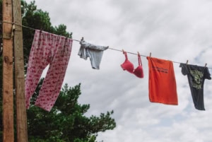 Photographie de corde à linge portant un pantalon de pyjama rose à motif floral kitsch, un T-shirt gris uni, un soutien gorge rose, un T-shirt orange avec l'inscription "cowboys fringants", et un T-Shirt noir décoré . Vue en contreplongée avec le ciel comme arrière plan.