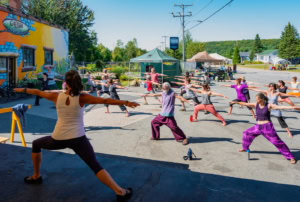 Photographie d'une séance de yoga avec des participants de tous les âges sur une petite placette d'un village au Québec, à côté du "garage de la culture"