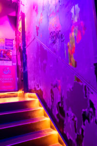 Vieil escalier aux murs usés et tagués, éclairé par des néons violets et jaunes, à Melbourne.